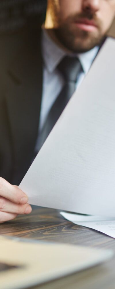 Portrait of modern bearded businessman holding papers in hands, reading and analyzing contract documentation at desk with laptop
